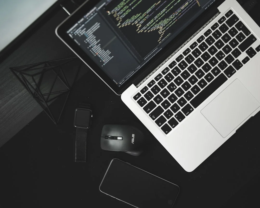 A mobile phone rests atop a wooden surface beside an out-of-focus laptop within a warm indoor environment.